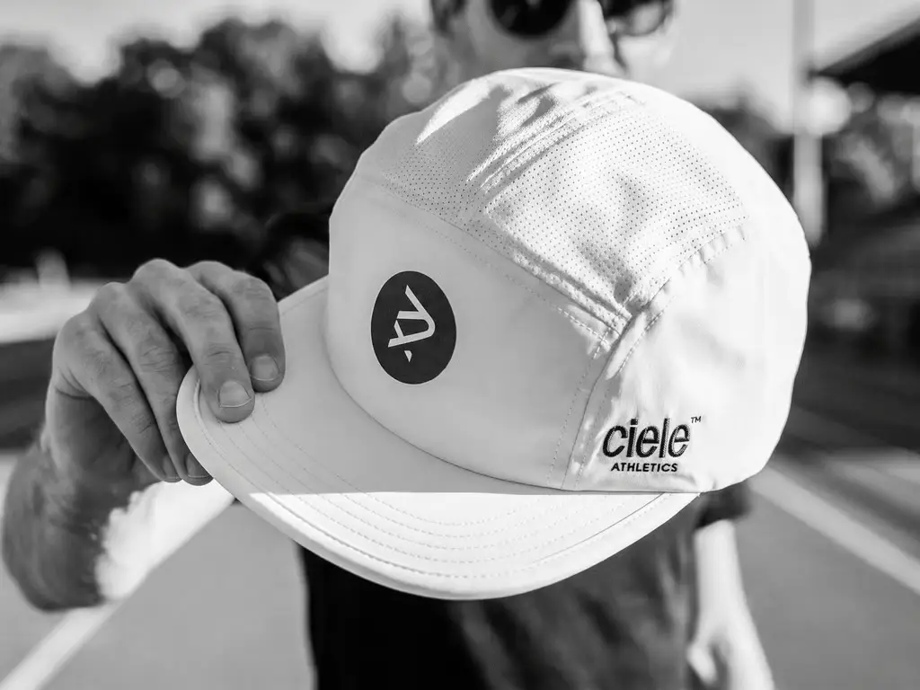 Casquette de course Sexy Pace Collective blanche, tenue à la main sur une piste