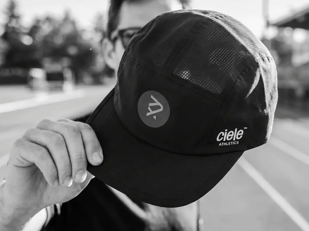 Casquette de course Sexy Pace Collective noire, tenue à la main sur une piste